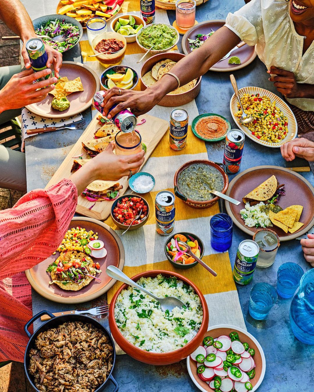 Overhead shot of a vibrant outdoor table laden with diverse Mexican dishes, drinks, and the hands of a group enjoying a shared meal.