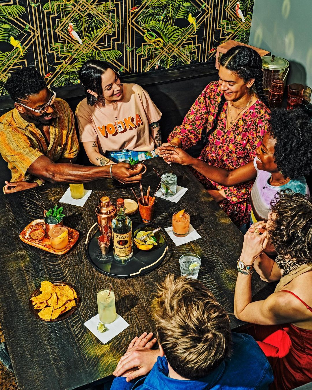 Six smiling people gathered around a dark wood table with drinks and appetizers. Tropical print wallpaper provides a vibrant backdrop.