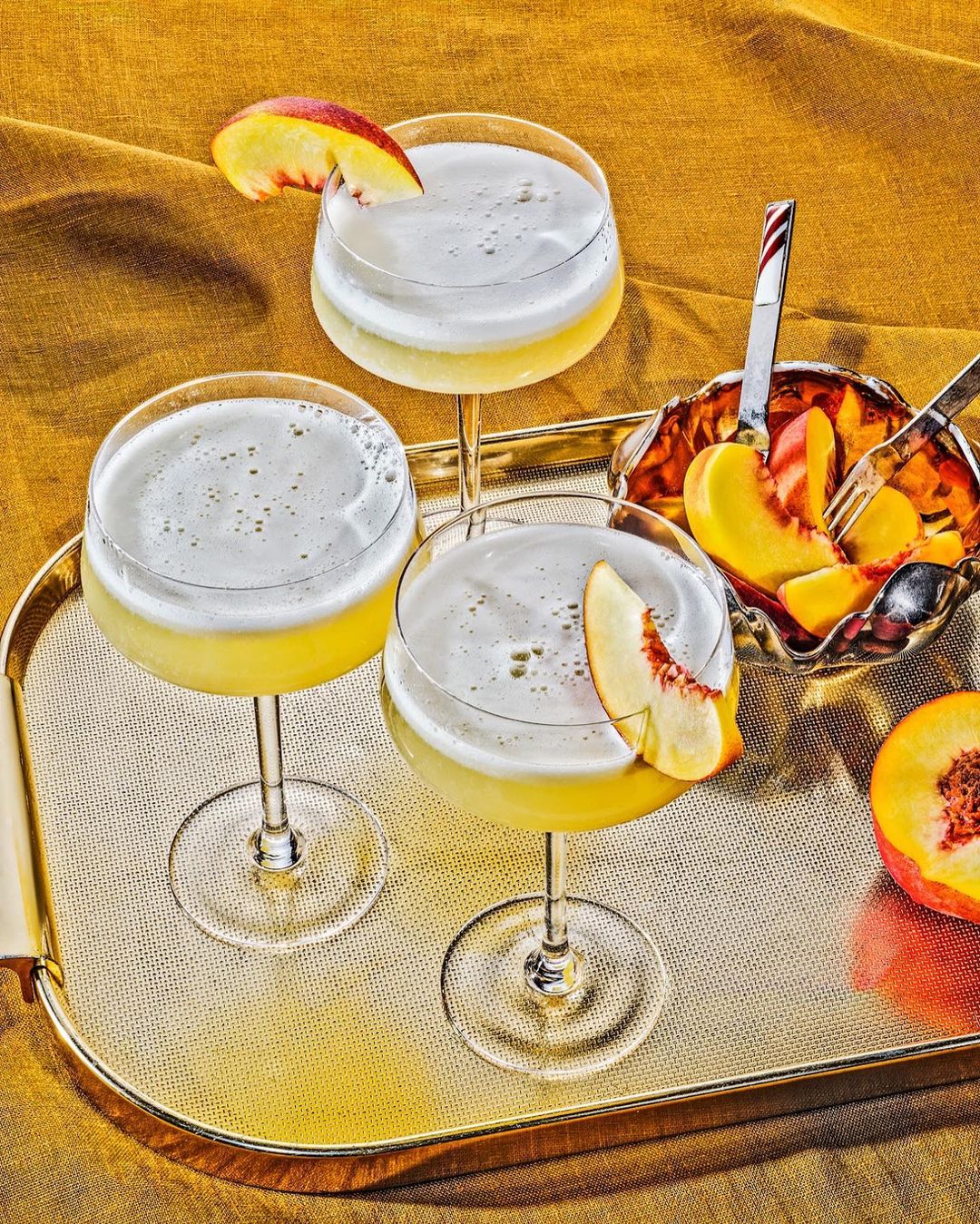 Three stem glasses of peach cocktails with foam and fruit slices on a gold tray over a textured yellow-gold fabric.
