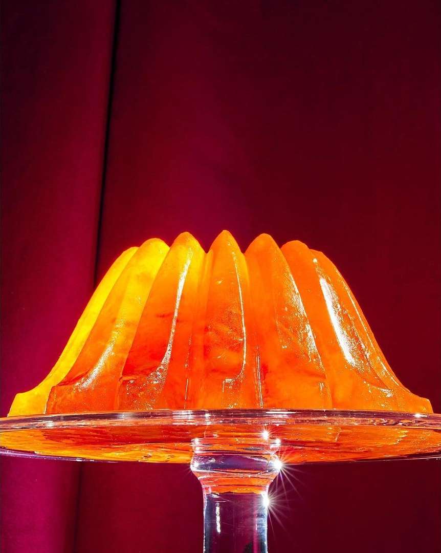 Translucent orange gelatin dessert in decorative mold on a stemmed glass serving platter in front of red fabric backdrop.