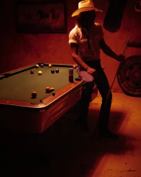 Man wearing a cowboy hat sitting on a pool table holding a cue stick in a warmly lit room by Johnny Autry.