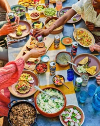 Overhead view of people gathering around a rustic outdoor dining table filled with Mexican food, tacos, and drinks.