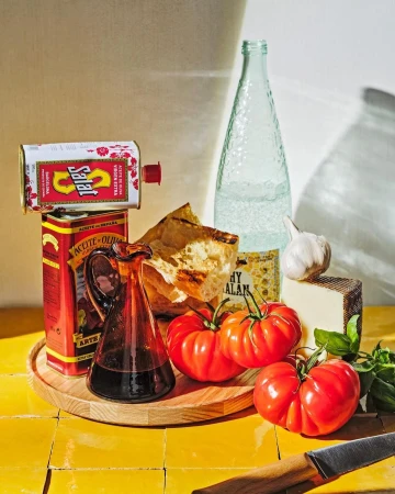 Still life food photography featuring olive oil tins, fresh tomatoes, bread, garlic, and a bottle of water on a yellow tiled surface.