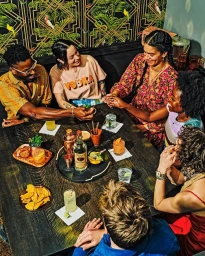 Group of six diverse friends gathered around a dark wooden table in a bar holding cocktails and snacks