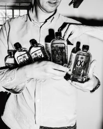 Monochrome medium shot of a man holding several glass bottles labeled XXX Sotol