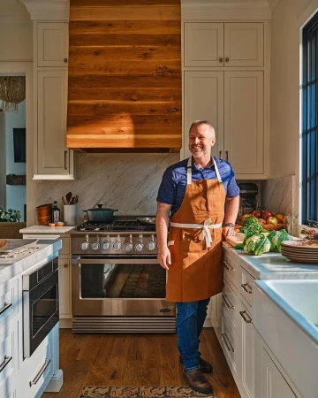 Man wearing a brown apron and blue shirt standing in a bright modern kitchen by a stainless steel stove