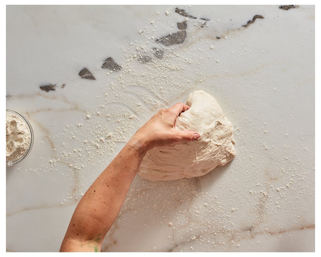 Close-up of an adult hand kneading a ball of dough on a white marble surface dusted with flour, with a bowl of flour visible.