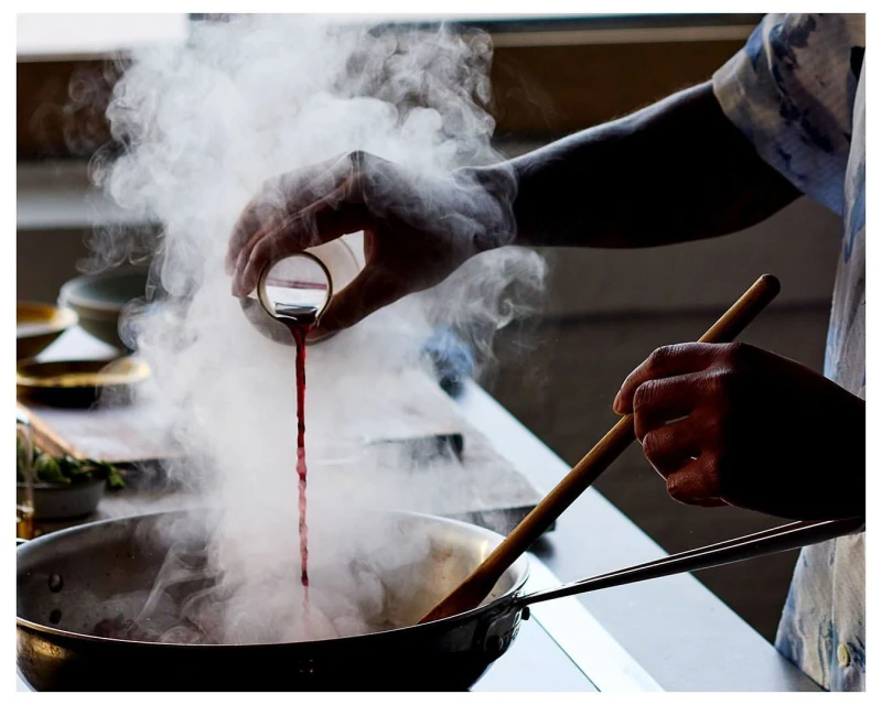An adult pours liquid into a skillet with a wooden spoon while cooking in an indoor kitchen setting, creating a steamy culinary scene.