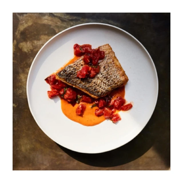 Overhead shot of a piece of cooked fish topped with diced tomatoes and herbs, served on a white plate with sauce.