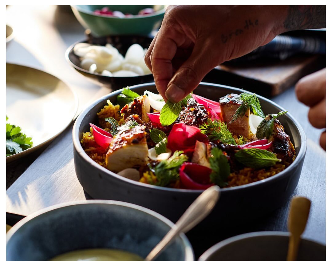 Close-up of a food bowl with garnished ingredients like greens and sliced onions, alongside a tattooed hand adding fresh mint.