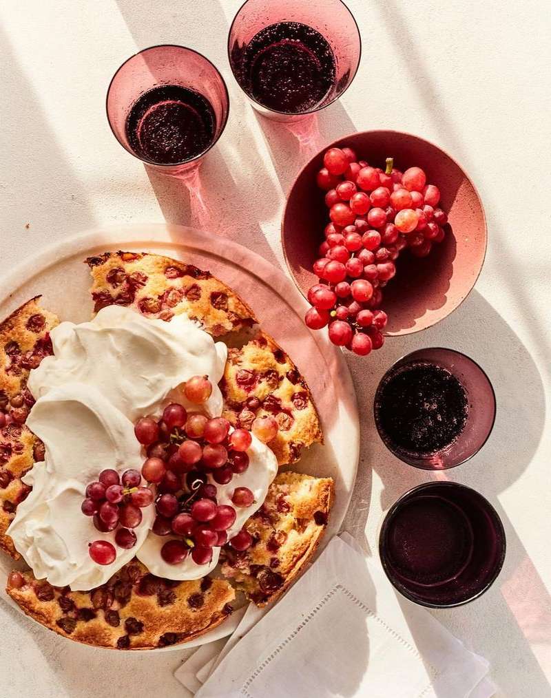 Overhead shot of grape cake topped with whipped cream, surrounded by bunches of grapes and stemware filled with a dark liquid.