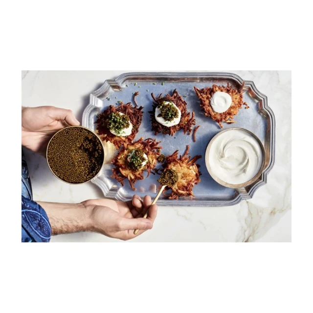 Elevated food presentation showcases potato latkes topped with cream and caviar on a silver tray, held by hands with a bowl and spoon.