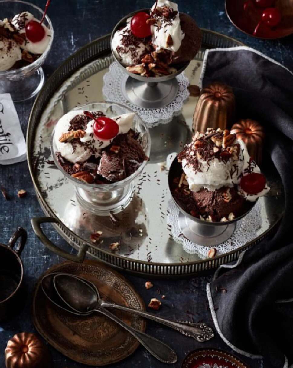 Overhead shot of four silver dessert cups filled with chocolate and vanilla ice cream topped with cherries and nuts on a tray.
