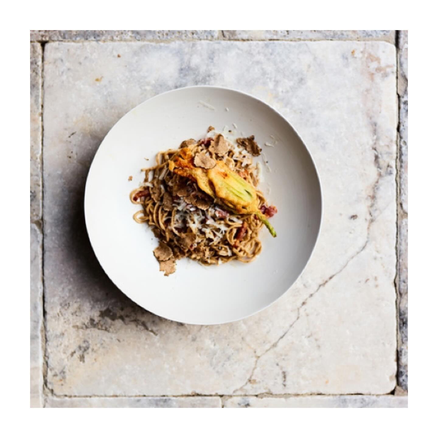 Overhead shot of a plate of spaghetti with a fried squash blossom and truffle shavings, set on a rustic marble tile surface.