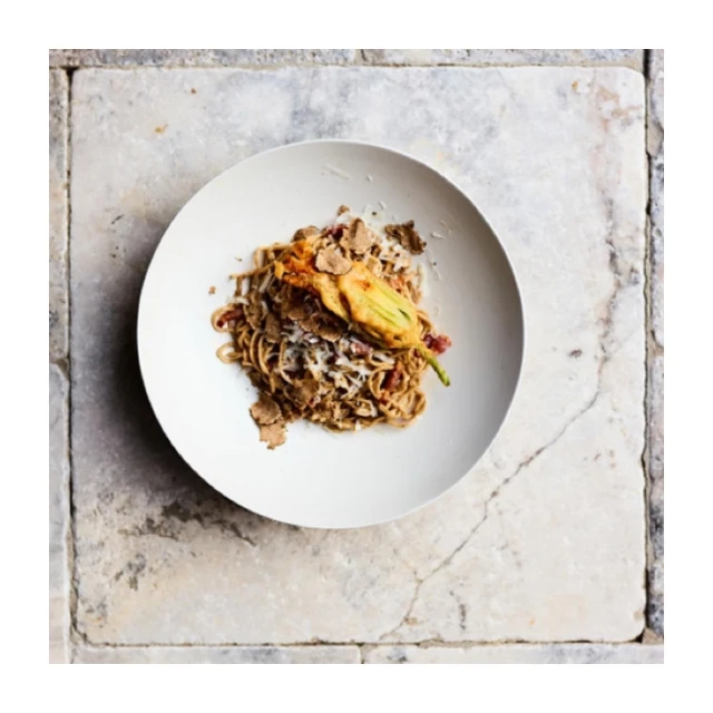 Overhead shot of a plate of spaghetti with a fried squash blossom and truffle shavings, set on a rustic marble tile surface.
