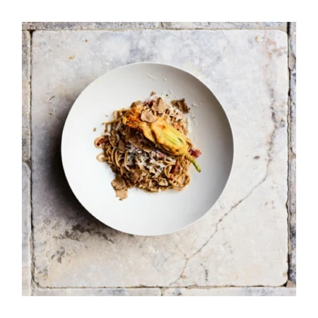 Overhead shot of a plate of spaghetti with a fried squash blossom and truffle shavings, set on a rustic marble tile surface.
