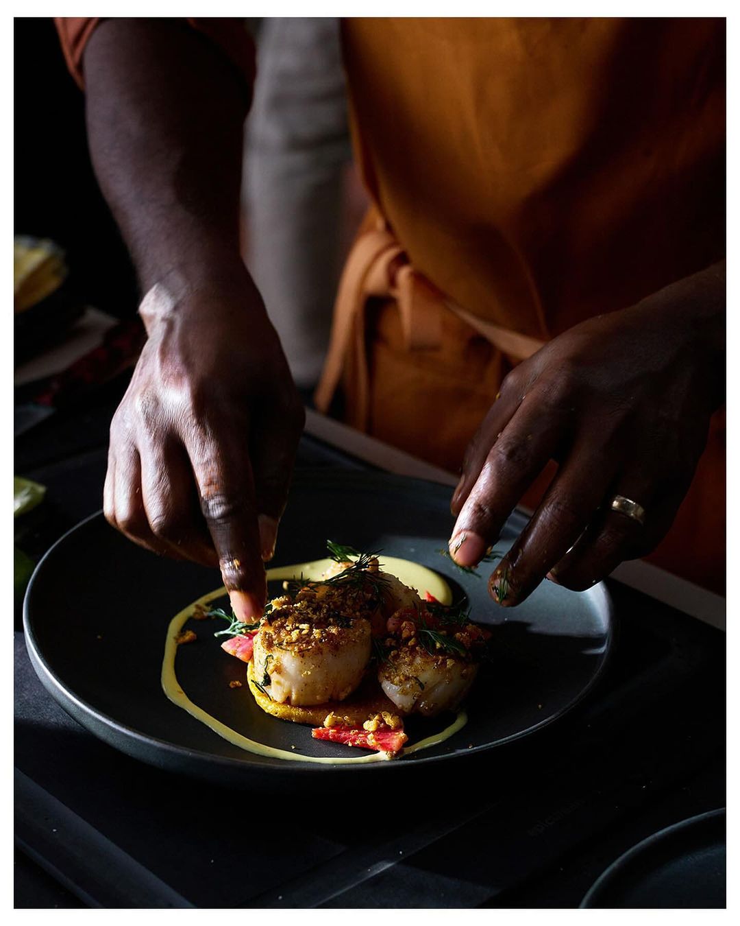 Two pan-seared scallops plated with a swirl of yellow sauce are being garnished by a chef wearing an orange apron in a dark setting.