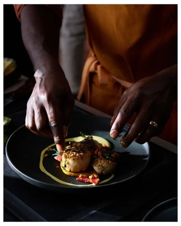 Two pan-seared scallops plated with a swirl of yellow sauce are being garnished by a chef wearing an orange apron in a dark setting.