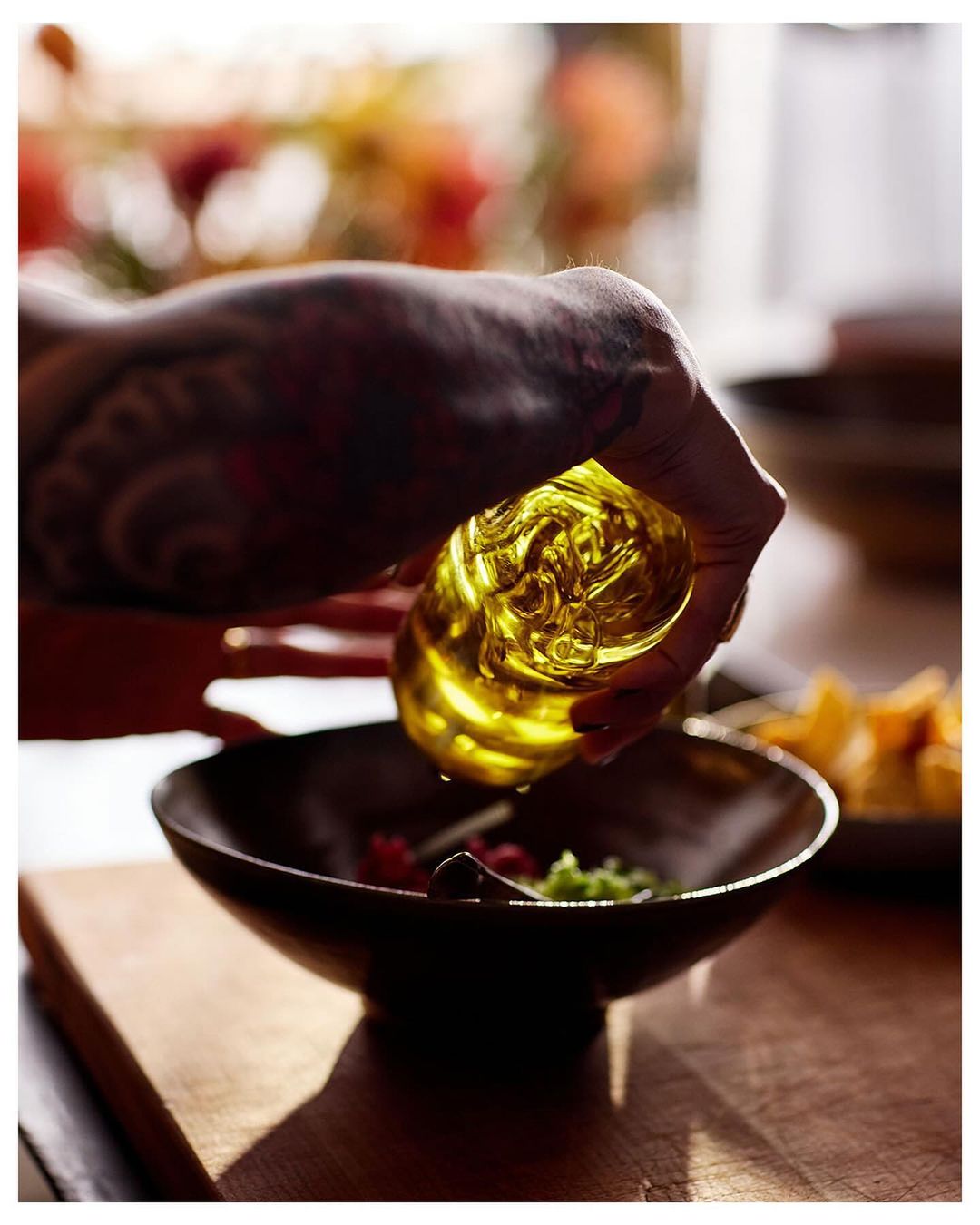 Person with tattooed arm pouring golden olive oil from glass bottle into bowl of salad on wooden table in natural light.