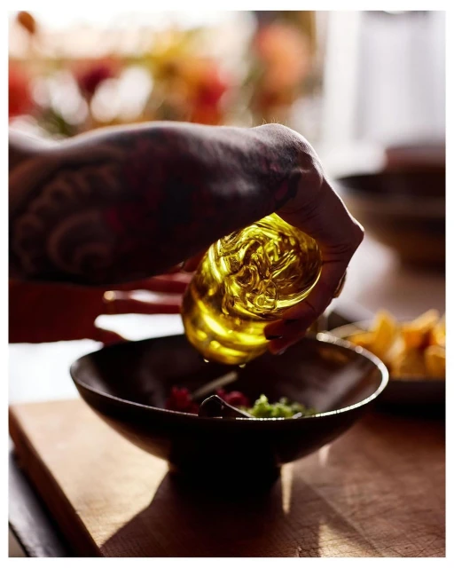 Person with tattooed arm pouring golden olive oil from glass bottle into bowl of salad on wooden table in natural light.