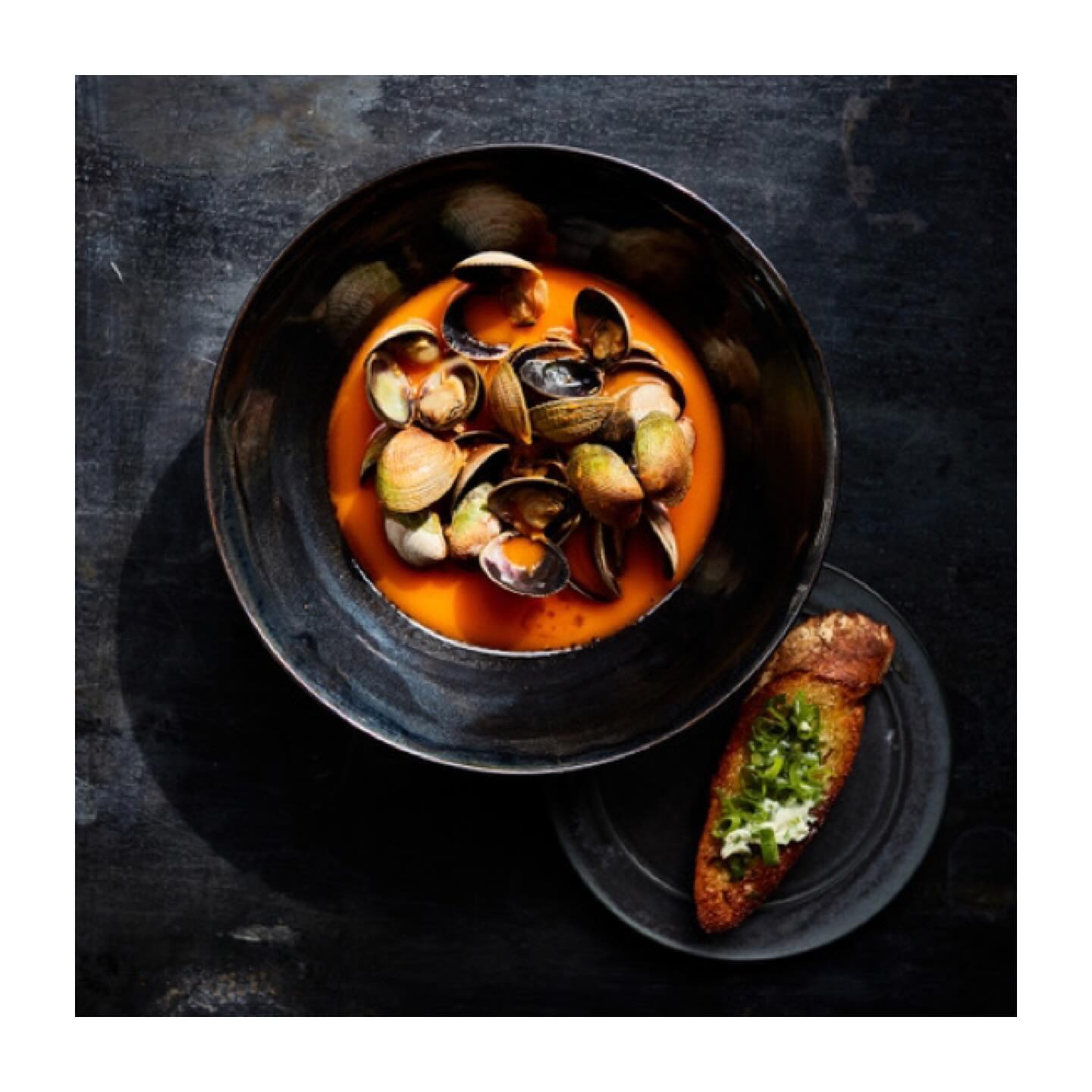Overhead shot of clam stew in a dark bowl with toasted bread on a small plate, set against a dark background, food photography.