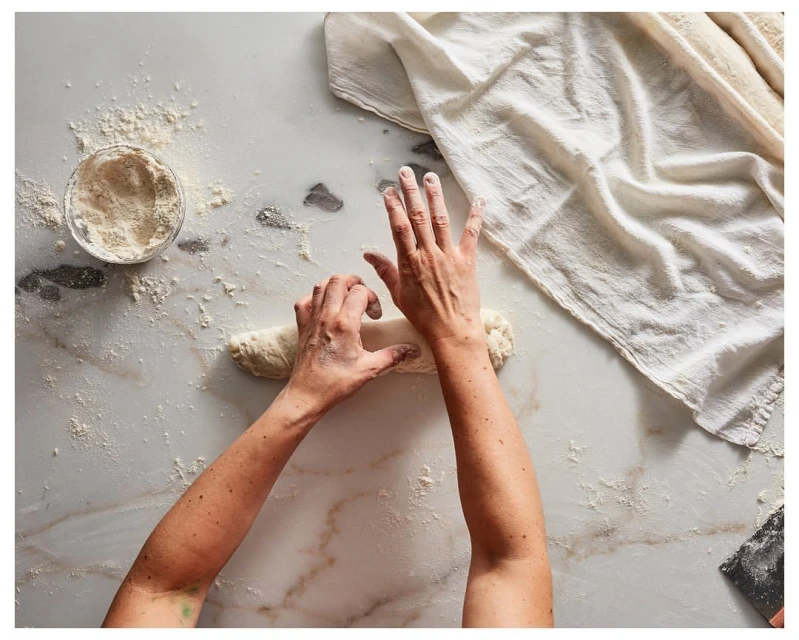 Overhead shot of hands kneading dough on a marble surface, with a bowl of flour and a white cloth in the background.