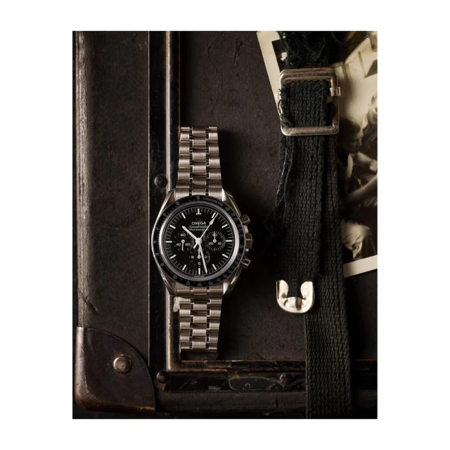 Silver Omega watch with black face on a metal band displayed atop a vintage brown suitcase with a black strap detail and old photograph.