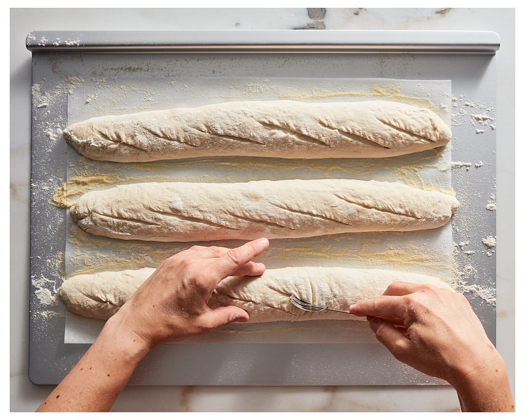 Three uncooked baguettes on baking sheet being scored by female hands with a fork, dusted with flour, prepared for baking.