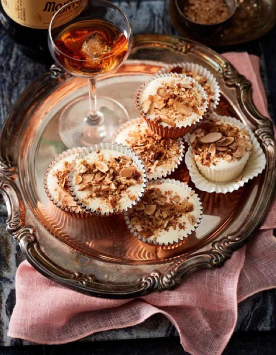 Silver tray containing a collection of cream-filled, nut-topped cupcakes next to a glass of amber liquid and a dark glass bottle.