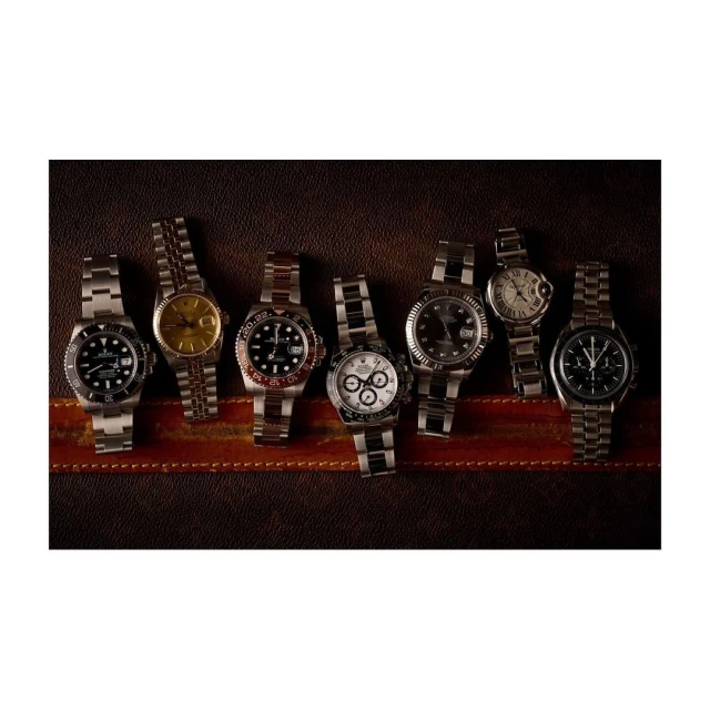 Seven silver-banded wristwatches of varying styles are displayed on a dark brown textured surface. The arrangement is clean and well-lit.