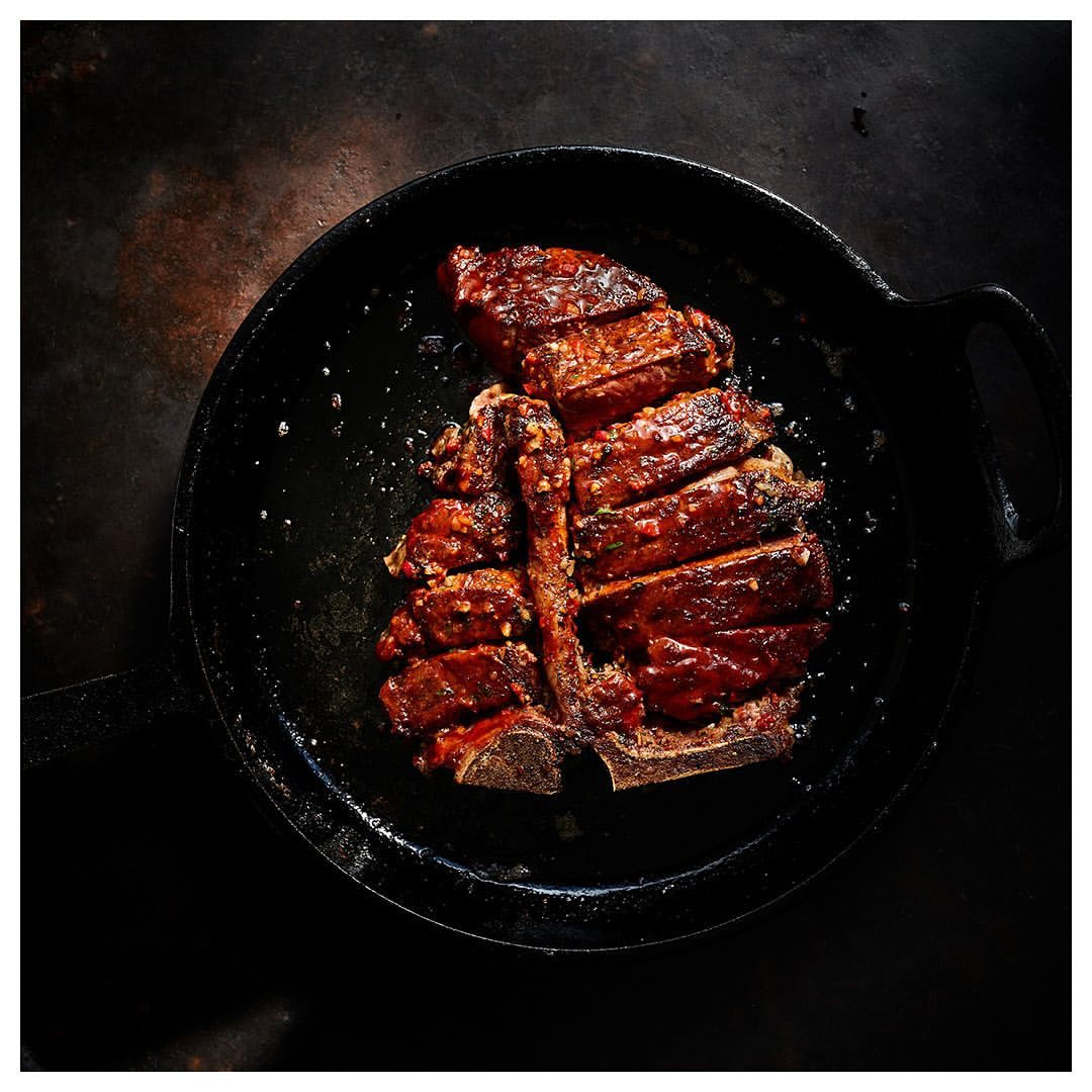 Overhead shot of a sliced, glazed T-bone steak served in a black cast iron skillet, displayed on a rustic dark background, food photography.