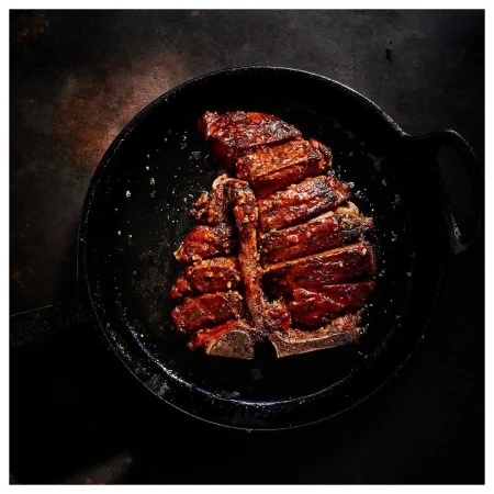 Overhead shot of a sliced, glazed T-bone steak served in a black cast iron skillet, displayed on a rustic dark background, food photography.