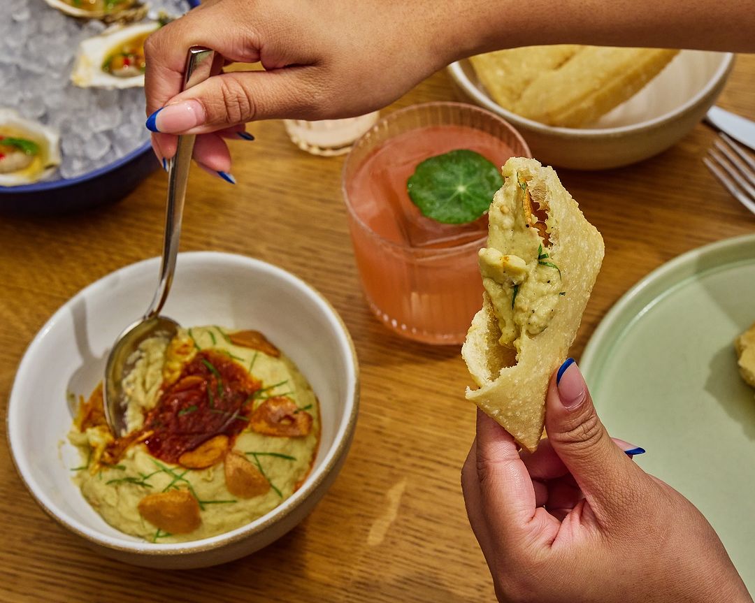 Close-up of hands holding a filled pastry, with a bowl of dip, a drink, and oysters on a wooden table. Food and beverage scene.