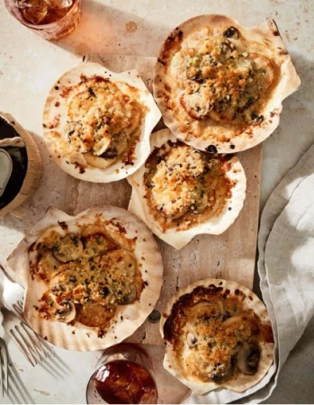 Overhead shot of five scallop shells filled with a creamy mixture and topped with browned breadcrumbs, served on a wooden board.