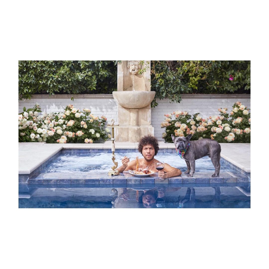 Adult male and French bulldog dog in a pool with water fountain, plate of food, and red wine. Summer daytime outdoor scene.