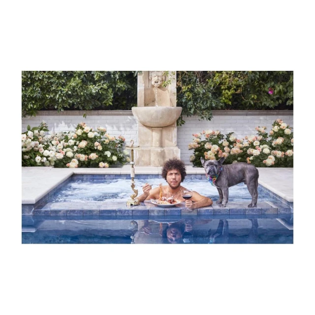 Adult male and French bulldog dog in a pool with water fountain, plate of food, and red wine. Summer daytime outdoor scene.