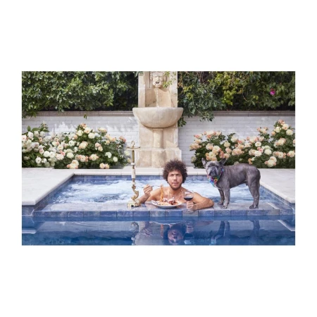 Adult male and French bulldog dog in a pool with water fountain, plate of food, and red wine. Summer daytime outdoor scene.