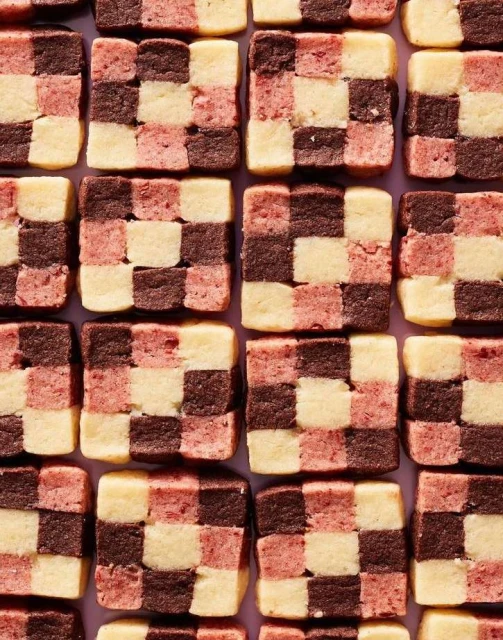 Overhead shot of a checkerboard pattern made of square cookies in brown, light pink, and cream arranged on a light purple surface.