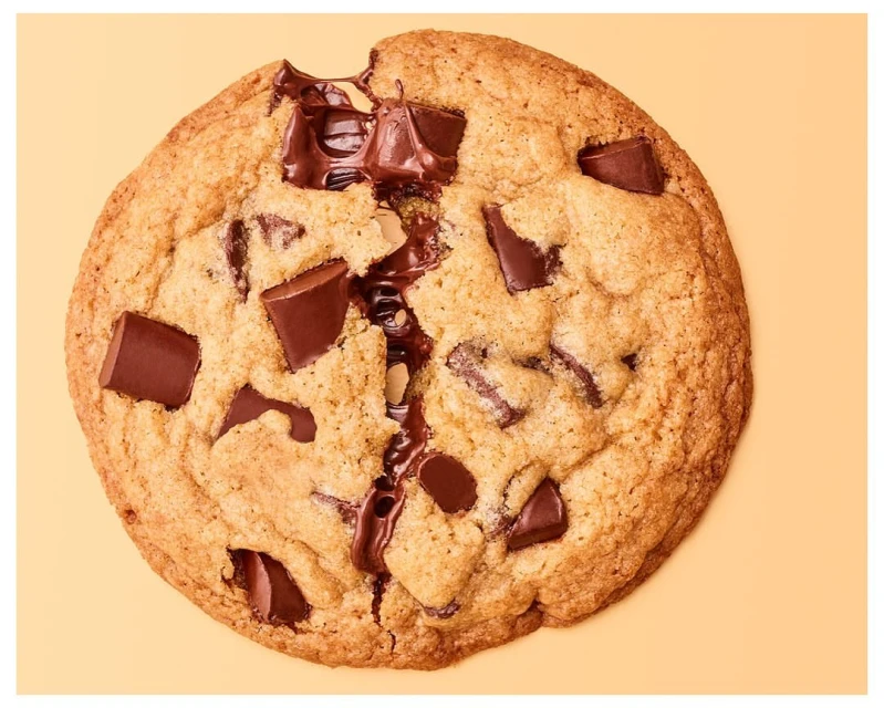 Top-down shot of a chocolate chip cookie with melted chocolate chunks on a light orange background, perfect for food and dessert-themed projects.