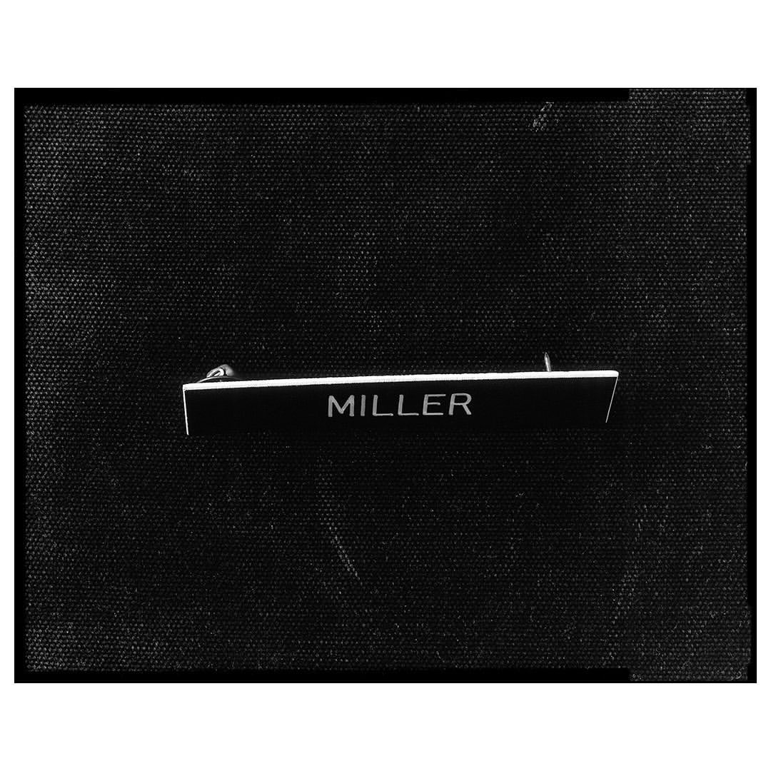 Close-up of a black name tag with the word 'MILLER' in white letters, attached to black fabric in a studio setting.