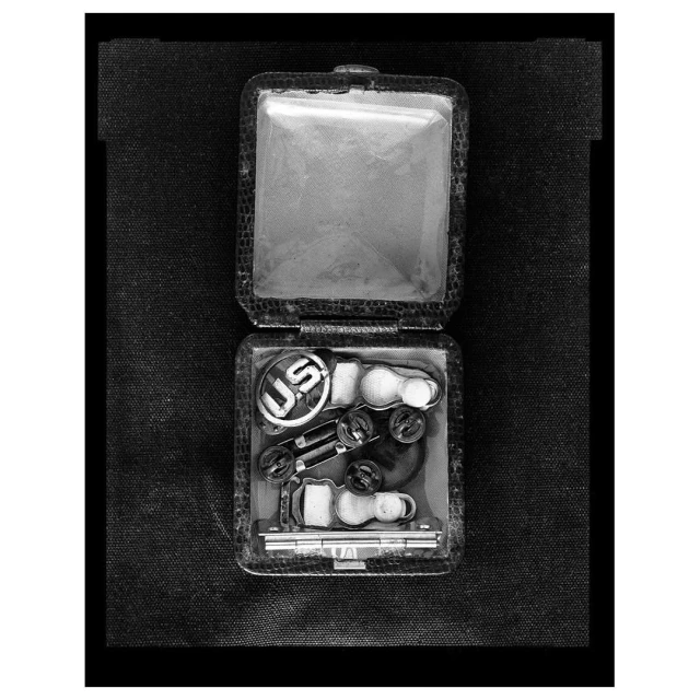 Black and white studio shot of an opened box containing U.S. military insignia, badges, and pins against a dark fabric backdrop.
