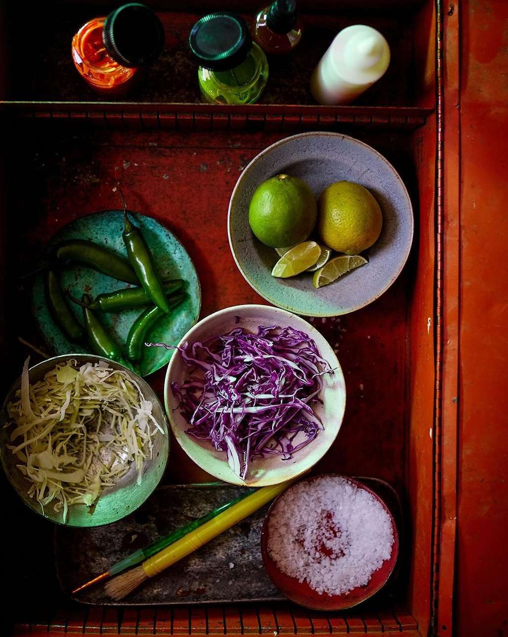Overhead shot of ingredients for Mexican cooking: limes, cabbage, chilis, and salt, all in bowls on a red surface with bottles in the background.