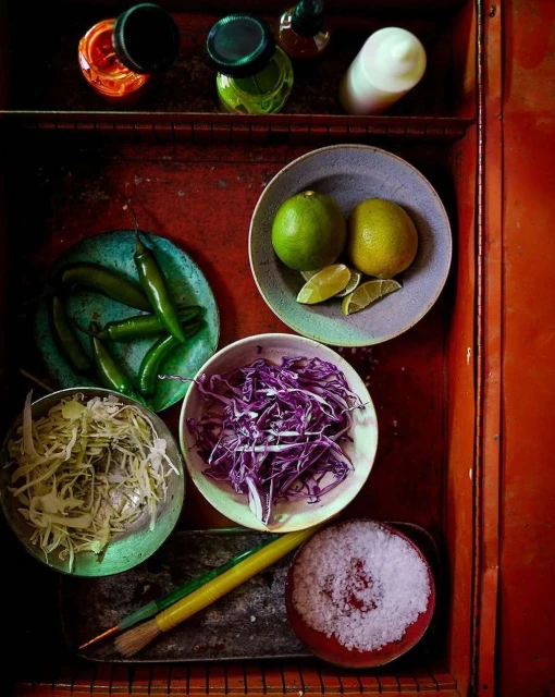 Overhead shot of ingredients for Mexican cooking: limes, cabbage, chilis, and salt, all in bowls on a red surface with bottles in the background.