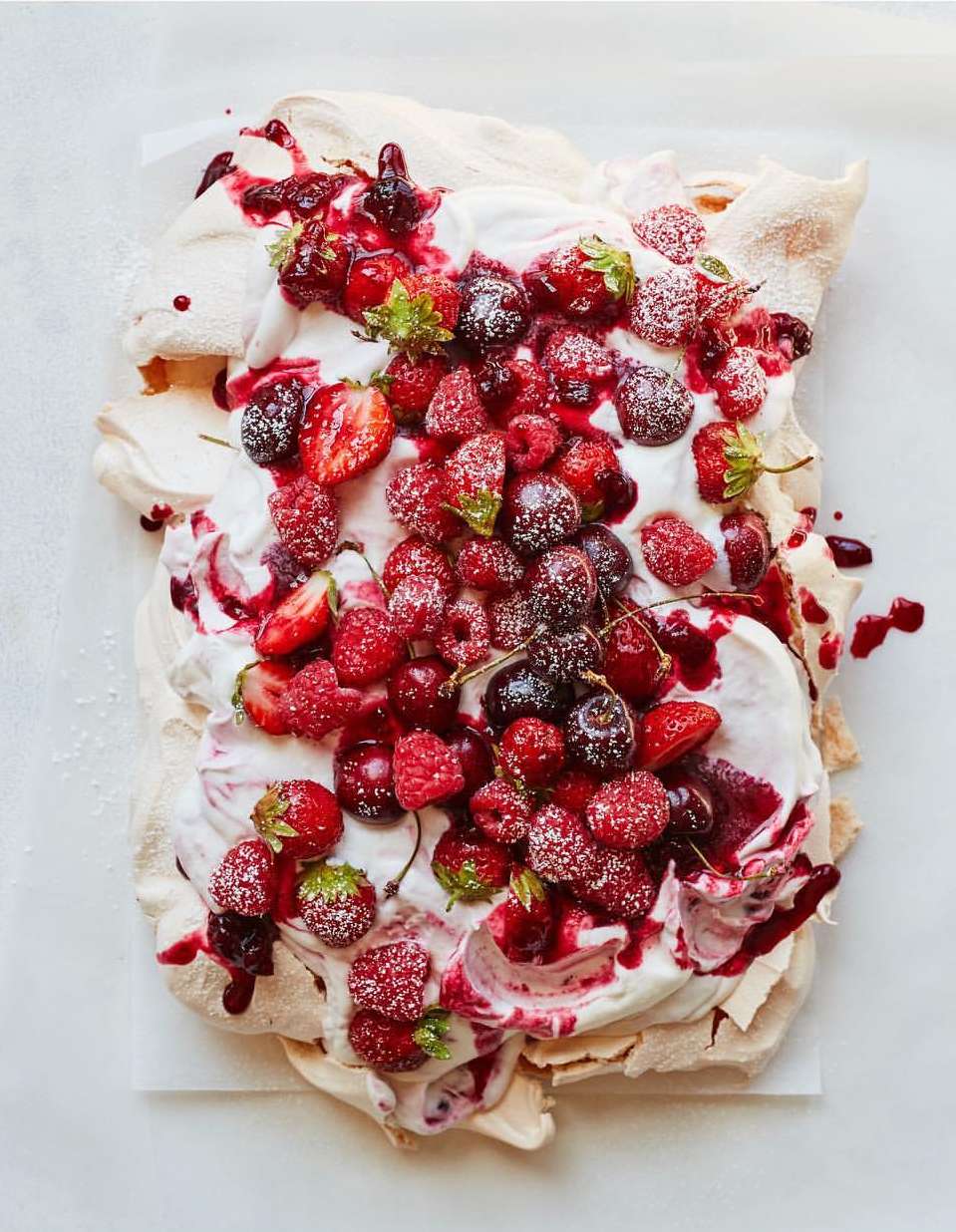 Overhead shot of a meringue dessert topped with whipped cream, strawberries, raspberries, cherries, and a red berry sauce.