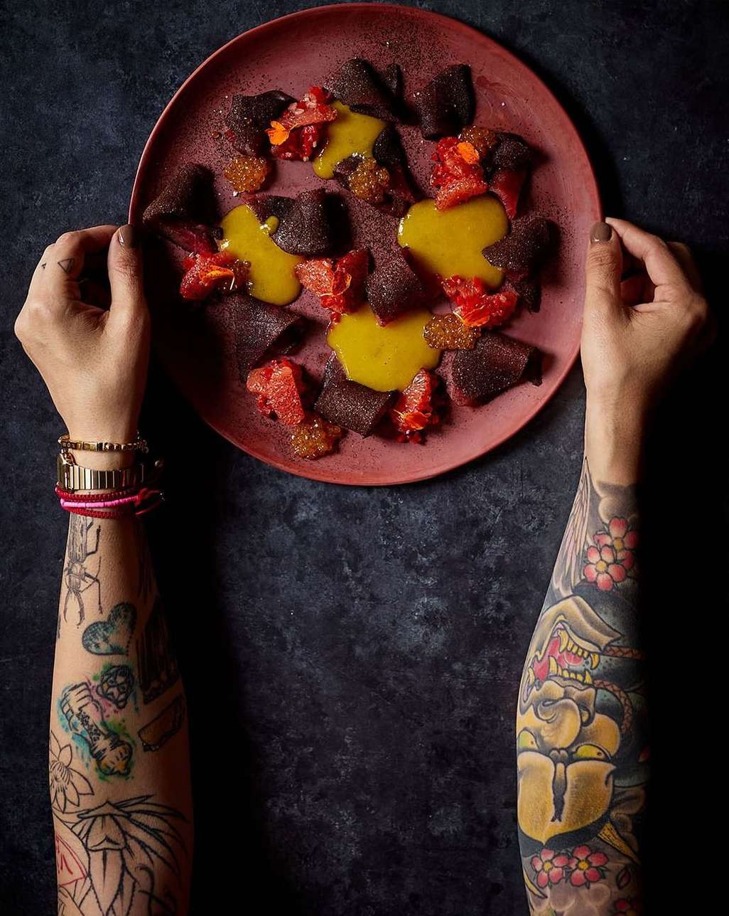 Overhead shot of a culinary food art dish on a plate held by two hands with arm tattoos, featuring beets, sauce, and citrus fruit.