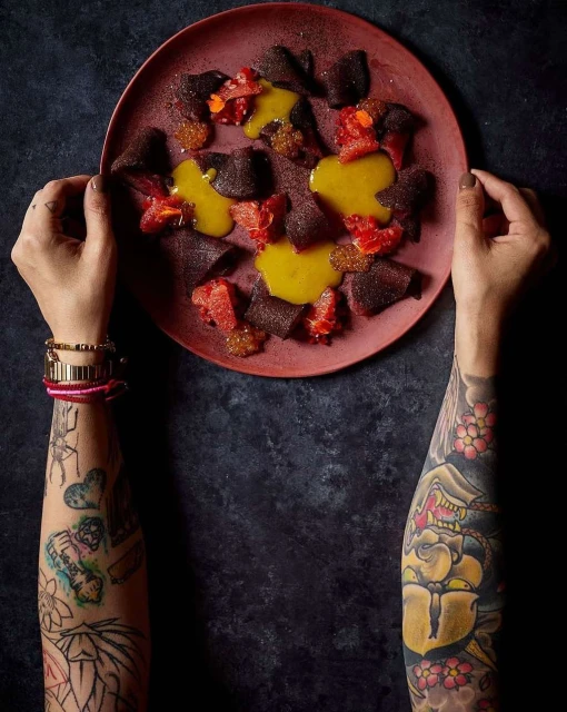 Overhead shot of a culinary food art dish on a plate held by two hands with arm tattoos, featuring beets, sauce, and citrus fruit.
