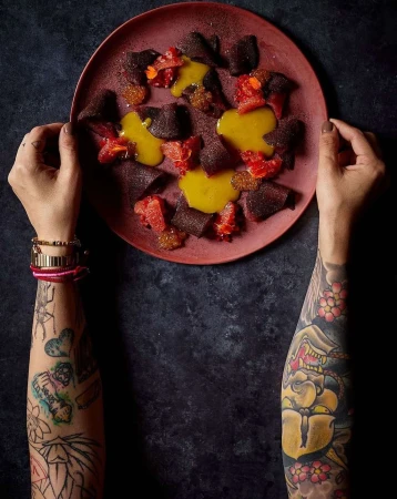 Overhead shot of a culinary food art dish on a plate held by two hands with arm tattoos, featuring beets, sauce, and citrus fruit.