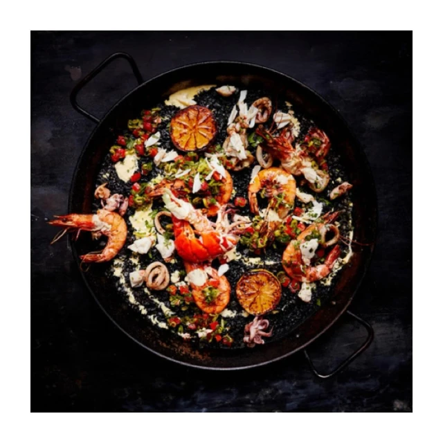 Overhead shot of a paella pan filled with seafood and lemons on a black surface creating a visually appealing culinary presentation.