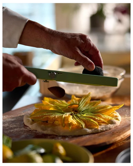 Chef grates black truffle over squash blossom pizza on a wooden board, showcasing culinary artistry and fresh ingredients in a warm indoor kitchen setting.