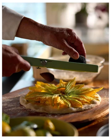 Chef grates black truffle over squash blossom pizza on a wooden board, showcasing culinary artistry and fresh ingredients in a warm indoor kitchen setting.